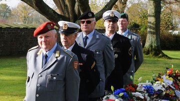 Fünf uniformierte Männer stehen gestaffelt hintereinander vor großen Bäumen. Neben ihnen sieht man bunte Blumenkränze.