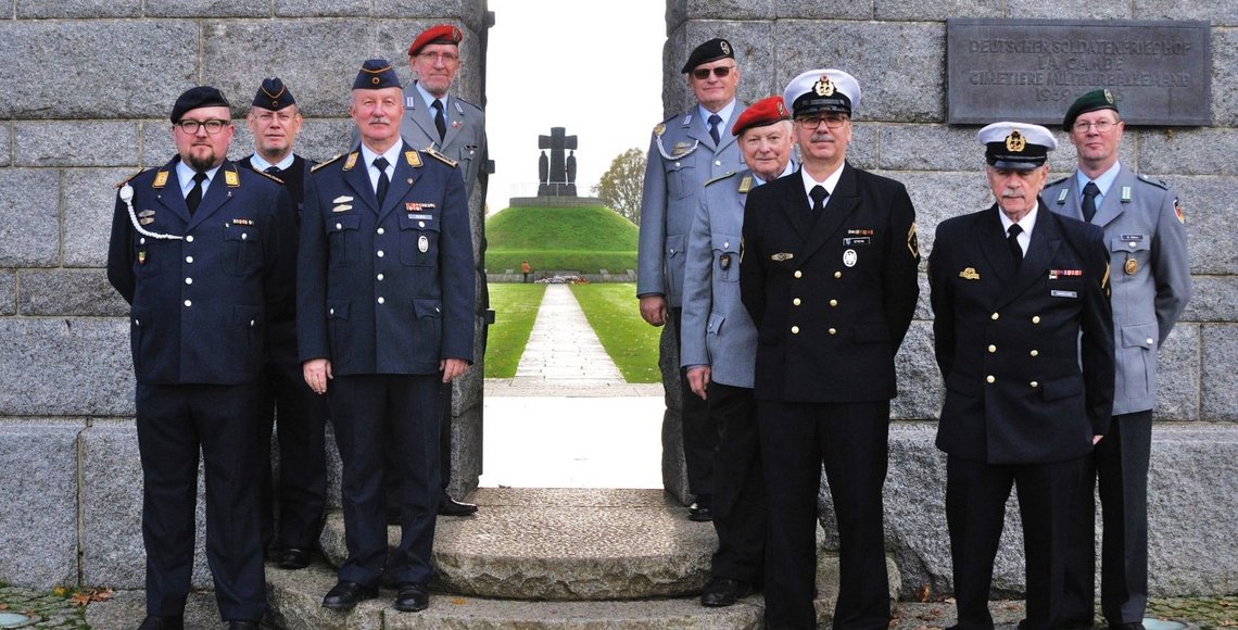 9 Männer in den Uniformen unterschiedlicher Waffengattungen stehen vor einer Mauer, rechts und links eines Tores. Durch das Tor sieht man den Tumulus (Hügel mit Denkmal) der Kriegsgräberstätte La Cambe.