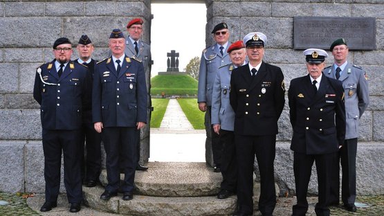 9 Männer in den Uniformen unterschiedlicher Waffengattungen stehen vor einer Mauer, rechts und links eines Tores. Durch das Tor sieht man den Tumulus (Hügel mit Denkmal) der Kriegsgräberstätte La Cambe.