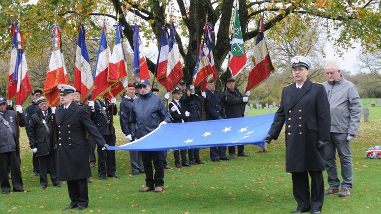 Mehrere uniformierte Männer stehen auf einer Rasenfläche unter einem großen Baum. Vier von ihnen halten die Europaflagge aufgespannt zwischen sich. Dahinter halten andere Männer französische Fahnen. Im Hintergrund sieht man Grabseine von Kriegsgräbern.