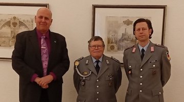 Drei Herren, darunter zwei in Uniform, stehen nebeneinander und lächeln in die Kamera.