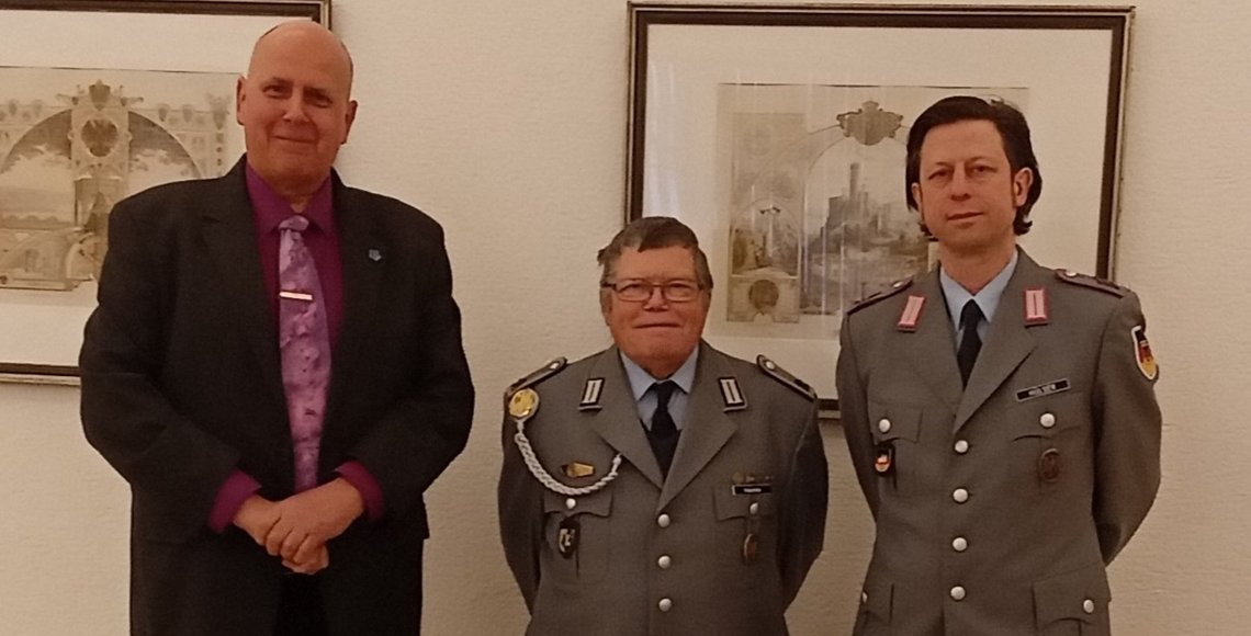Drei Herren, darunter zwei in Uniform, stehen nebeneinander und lächeln in die Kamera.