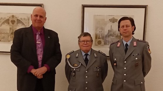 Drei Herren, darunter zwei in Uniform, stehen nebeneinander und lächeln in die Kamera.