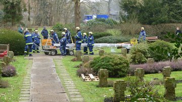 Man sieht mehrere Personen in blauen Uniformen und mit gelben Helmen, die das schwere Kreuz über den Friedhof tragen.